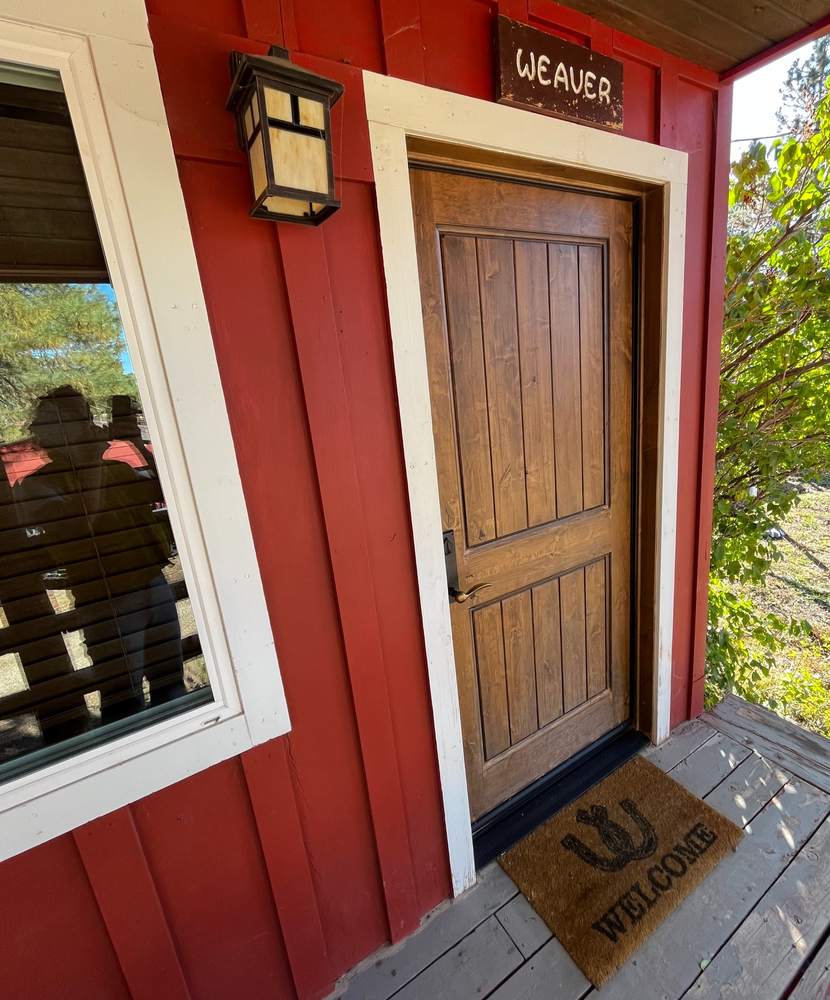 The wooden front door of a red cabin beneath a “Weaver” name sign and a porch lantern