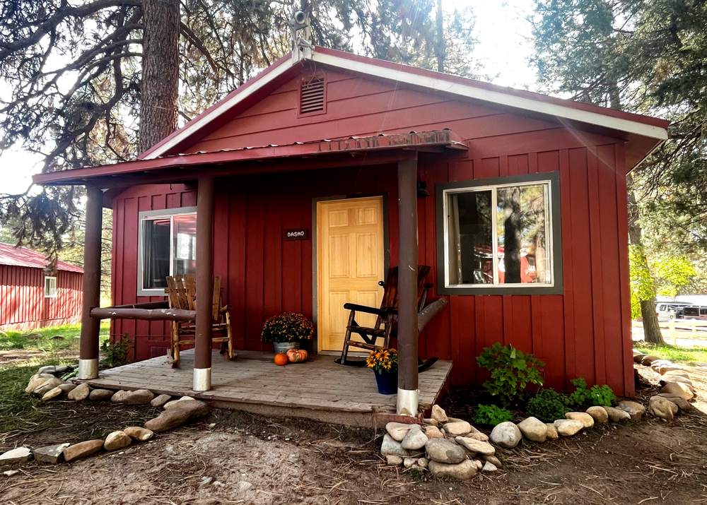 A red board-and-batten cabin with a covered porch and rocking chairs, under ponderosa pines